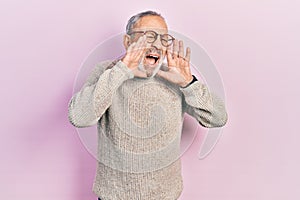 Handsome senior man with beard wearing casual sweater and glasses shouting angry out loud with hands over mouth