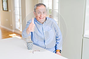 Handsome senior man drinking a cup of coffee at home doing happy thumbs up gesture with hand