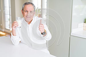 Handsome senior man drinking a glass of fresh milk at breakfast happy with big smile doing ok sign, thumb up with fingers,