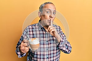 Handsome senior man with grey hair drinking a cup coffee asking to be quiet with finger on lips