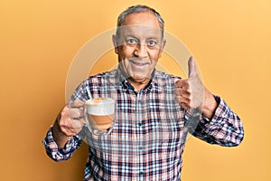 Handsome senior man with grey hair drinking a cup coffee doing happy thumbs up gesture with hand