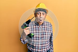 Handsome senior man with grey hair holding electric screwdriver wearing hardhat scared and amazed with open mouth for surprise,