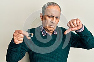 Handsome senior man with grey hair holding medical hearing aid with angry face, negative sign showing dislike with thumbs down,