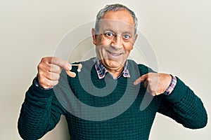 Handsome senior man with grey hair holding medical hearing aid pointing finger to one self smiling happy and proud
