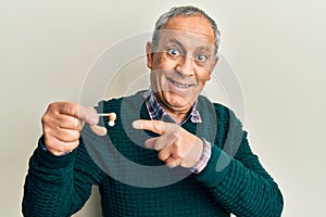 Handsome senior man with grey hair holding medical hearing aid smiling happy pointing with hand and finger