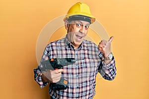 Handsome senior man with grey hair holding screwdriver wearing hardhat pointing thumb up to the side smiling happy with open mouth