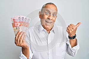 Handsome senior man holding 100 hong kong dollars banknotes pointing thumb up to the side smiling happy with open mouth