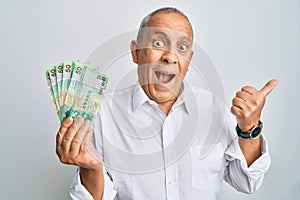 Handsome senior man holding 50 hong kong dollars banknotes pointing thumb up to the side smiling happy with open mouth