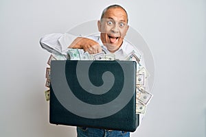 Handsome senior man holding briefcase full of dollars sticking tongue out happy with funny expression