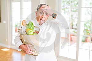 Handsome senior man holding a paper bag of fresh groceries from the supermarket stressed with hand on head, shocked with shame and