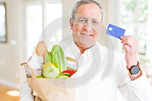 Handsome senior man holding paper bag full of fresh groceries and showing credit card as payment