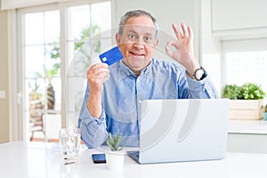 Handsome senior man shopping online using credit card and laptop at home doing ok sign with fingers, excellent symbol