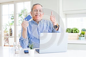 Handsome senior man shopping online using credit card and laptop at home happy with big smile doing ok sign, thumb up with