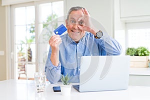 Handsome senior man shopping online using credit card and laptop at home with happy face smiling doing ok sign with hand on eye
