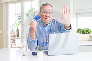 Handsome senior man shopping online using credit card and laptop at home with open hand doing stop sign with serious and confident