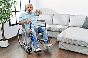 Handsome senior man sitting on wheelchair at the living room looking unhappy and angry showing rejection and negative with thumbs