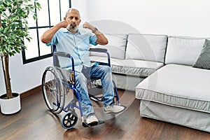 Handsome senior man sitting on wheelchair at the living room showing arms muscles smiling proud