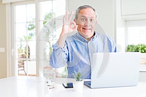 Handsome senior man using computer laptop working on internet doing ok sign with fingers, excellent symbol