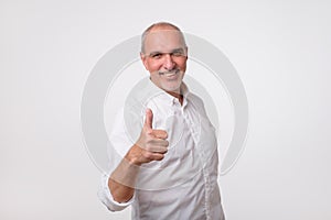 Handsome senior man in white shirt shows thumb up. He is satisfied with his health and financial conditio