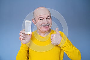 Handsome senior man in yellow sweater shows thumb up and phone screen