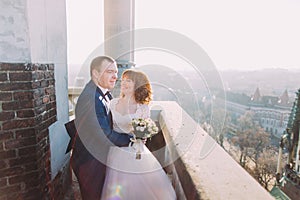Handsome sensual groom hugging newlywed bride from behind at old castle balcony with city background