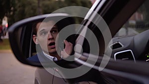 Handsome serious business man in suit and white shirt talking by mobile phone in the car. Reflection in the car`s door