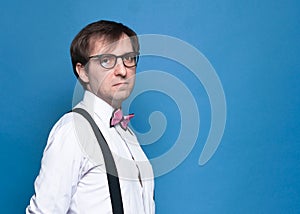 Handsome serious man in shirt, suspender, pink bow tie and glasses standing and looking at camera on blue background