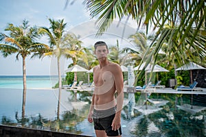 Handsome sexy young man standing near infinity swimming pool at the tropical island
