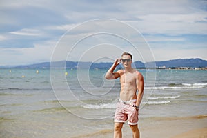 Handsome shirtless athletic young guy walking on the beach by the waterline. Young muscular man with perfect body in