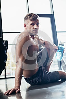 handsome shirtless sportsman sitting on floor in gym and holding sport bottle, looking photo