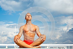 Handsome Shirtless Young Man During Meditation or Doing an Outdoor Yoga Exercise Sitting against the blue sky.