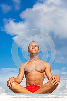 Handsome Shirtless Young Man During Meditation or Doing an Outdoor Yoga Exercise Sitting against the blue sky.