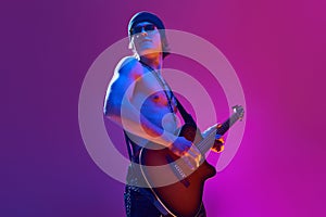 Handsome shirtless young man in sunglasses and hat playing electric guitar against pink background in neon light