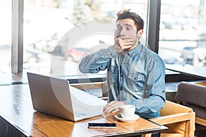 Handsome shocked scared man freelancer working on laptop, looking at screen covering mouth with palm