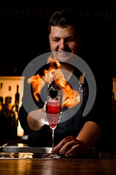 Handsome smiling bartender sets fire to the wine glass with cold tasty alcoholic drink