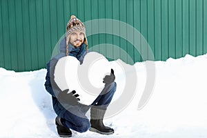 Handsome smiling bearded man in winter clothes holding a big heart made of snow, on green wall background. Declarations of love,