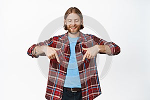 Handsome smiling blond man with beard, looking and pointing fingers down with pleased and happy face expression, showing