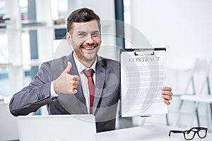 Handsome smiling businessman holding contract and showing thumb up in office