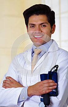 Handsome smiling doctor posing and looking at camera with a tensiometer in his hands with crossed arms, in office