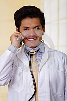 Handsome smiling doctor with an Stethoscope holding from his neck, using his celphone in office background