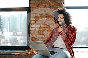 Handsome smiling egyptian man with beard sitting on floor against brick wall background and working in laptop