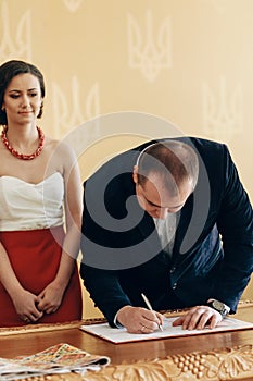 Handsome smiling groom in stylish suit signing marriage contract during wedding ceremony in registry office, newlywed family