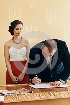 Handsome smiling groom in stylish suit signing marriage contract during wedding ceremony in registry office, newlywed family