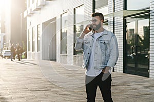 Handsome hipster businessman with beard, in denim jacket and trendy glasses walks around city and calls on mobile phone