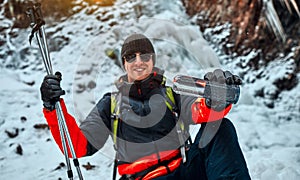 Handsome smiling man in assorted warm winter clothes and gloves holds a bottle of water and points to the camera