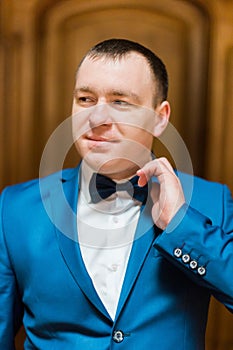 Handsome smiling man in blue suit fixin his bow tie in rich wooden interior