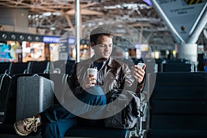 Handsome smiling man in casual wear holding luggage and messaging through his mobile phone while sitting at the hall of