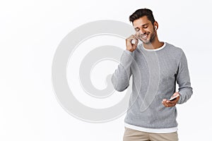 Handsome smiling relaxed guy with bristle in grey sweater, put on wireless earphones, listen music with new earbuds hold