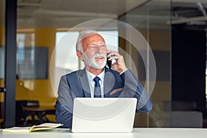 Handsome smiling senior man while sitting at his cozy workplace with laptop at home, retired male chatting with friends