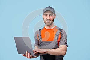 Handsome smiling workerman in uniform holding laptop and looking at camera on blue background